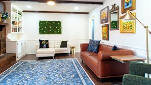 A living room with a blue floral rug, a white sofa, a brown leather sofa, and a green armchair. There is a bookshelf, a small side table, and a wall-mounted green plant. - Game Room + Colorful Comfort (Norman)