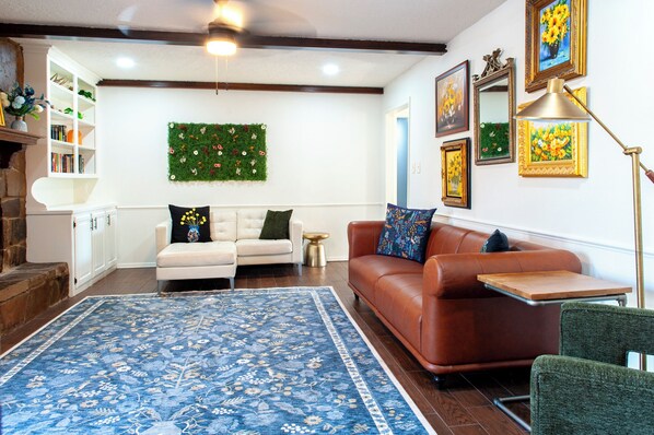 Ein Wohnzimmer mit blauem floralem Teppich, einem weißen Sofa, einem braunen Ledersofa und einem grünen Sessel. Es gibt ein Bücherregal, einen kleinen Beistelltisch und eine an der Wand befestigte grüne Pflanze.