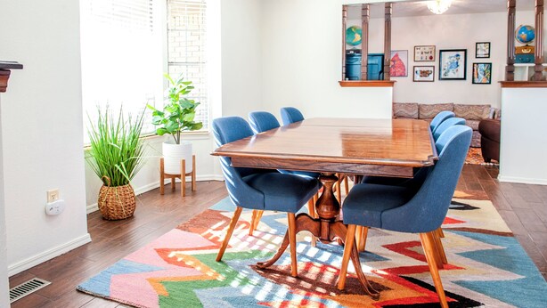 A dining area with a wooden table and blue upholstered chairs, a colorful rug, and a plant. - Game Room + Colorful Comfort (Norman)