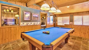 Ein Billardtisch in einem Raum mit Holzwänden und -decke, gerahmten Bildern und einem Steinkamin.