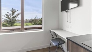 A room with a large window, a flat-screen TV mounted on the wall, and a white desk with a chair.