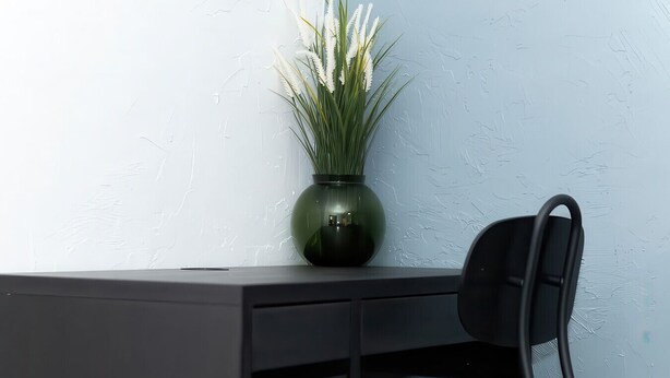 Een bureau met een stoel en een groene vaas met witte bloemen voor een structuurwand.