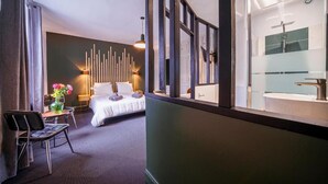 A hotel room with a bed, a chair, a table with a vase of flowers, a wall with vertical light fixtures, and a bathroom with a modern sink and mirror. - La Longère de Neuville (Neuville-de-Poitou)