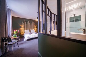 Quarto de hotel com cama, poltrona, mesa com um vaso de flores, parede com luminárias verticais e banheiro com pia e espelho modernos.