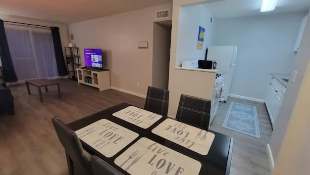 Ein Essbereich mit Tisch, gedeckt für vier Personen. Untersetzer mit der Aufschrift 'Live Love Eat'. Im Hintergrund steht ein Fernseher auf einem Schrank, und durch einen offenen Durchgang ist eine Küche zu sehen.
