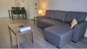 Ein Wohnzimmer mit einem grauen Ecksofa, einem Holztisch und einem Essbereich inklusive Tisch und Stühlen.