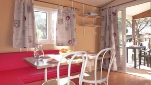 A dining area with a table, chairs, and a bench. There are curtains on the windows, and a small table with a pitcher and plates. - ZEcamping 4 étoiles locatif clim 24m2 4 pers (Hyères)