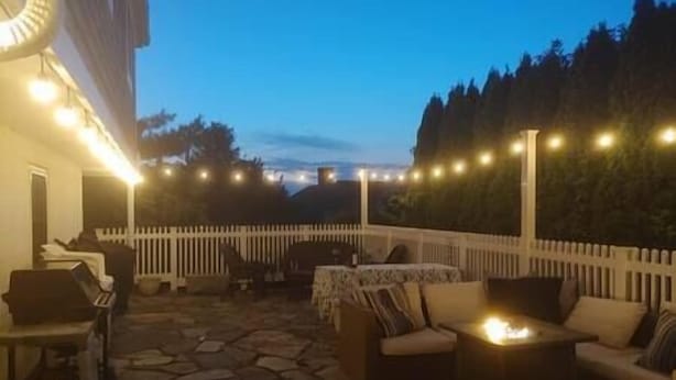 Un patio al aire libre con piso de piedra, cerca de piquetes blancos, luces colgantes y mobiliario exterior.