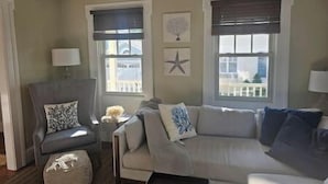 Una sala de estar con sofá, sillones y una mesita. Hay dos ventanas con persianas, una decoración con forma de estrella en la pared y una lámpara sobre una mesita auxiliar.