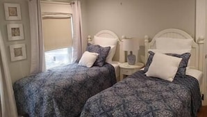 Un dormitorio con dos camas, una ventana con cortinas, una mesita de noche con lámpara y cuadros enmarcados en la pared.