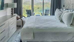 A bedroom with a large bed, a ceiling fan, a nightstand, a lamp, a dresser, and a view of mountains through large windows.