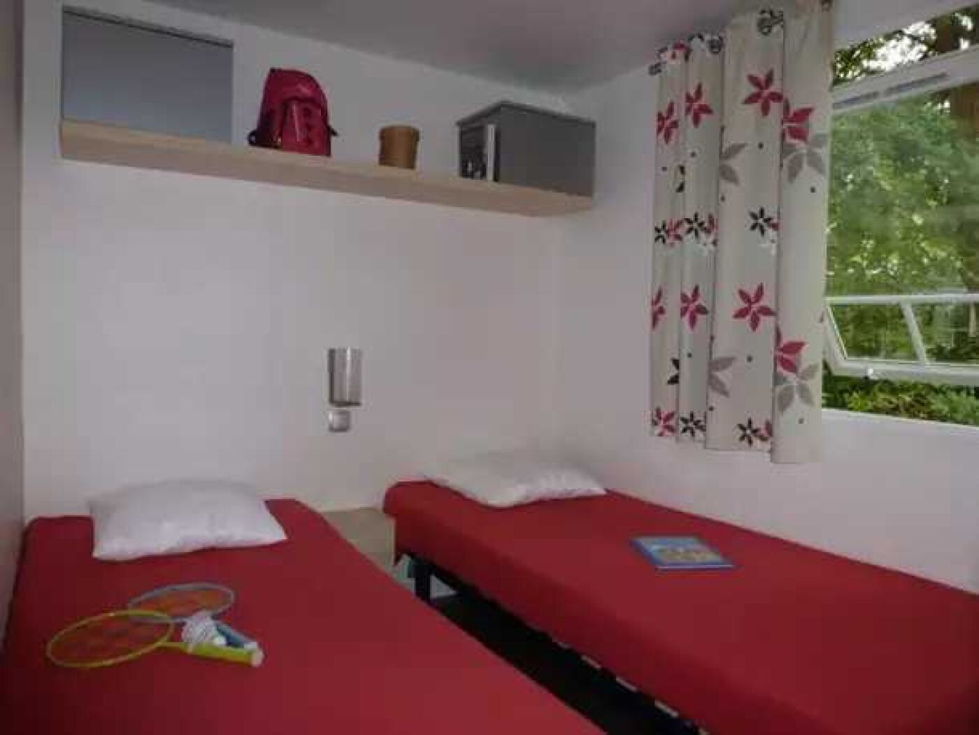 A room with two single beds, a small shelf with a red bag and a container, a window with floral curtains, and a tennis racket on one bed.