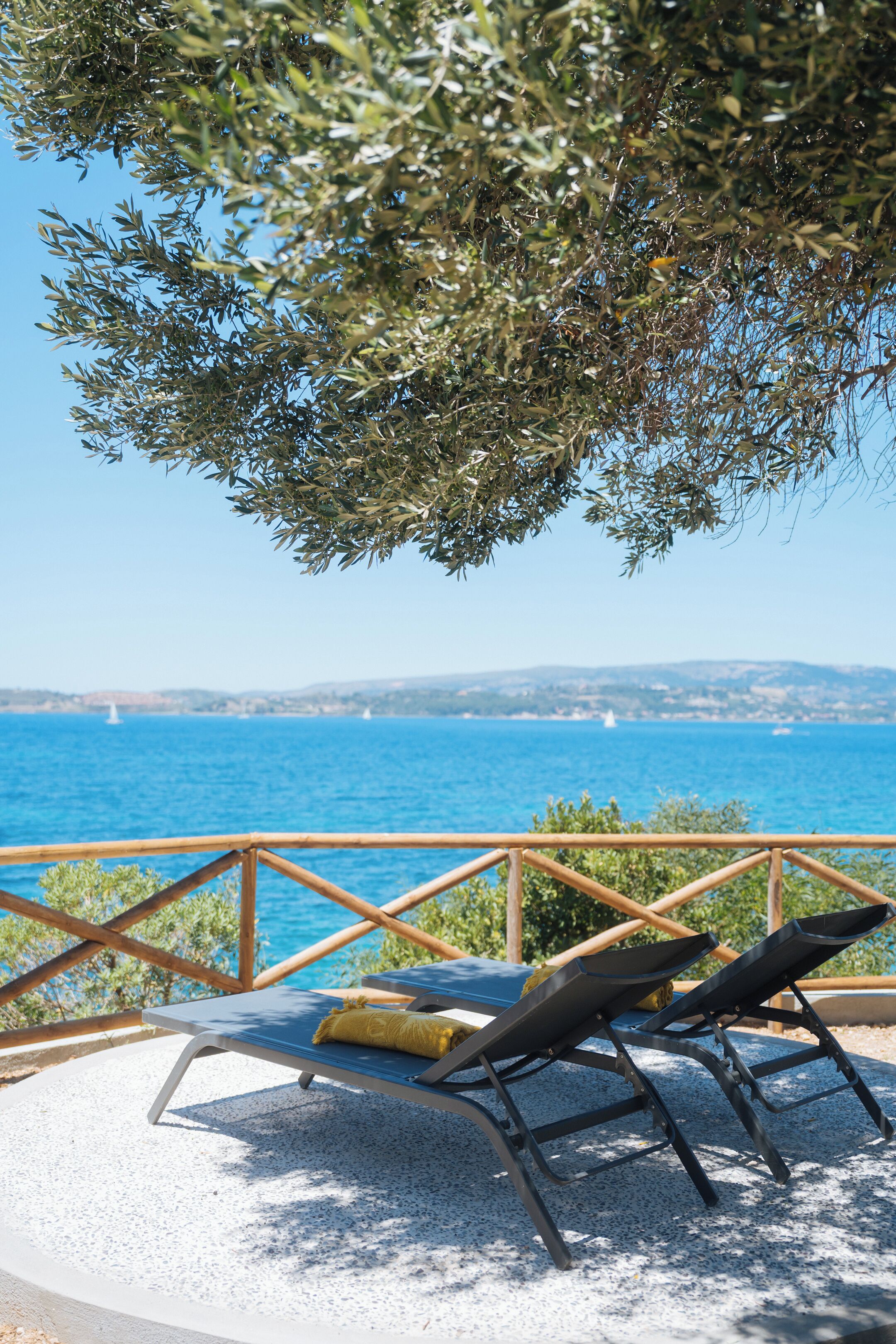 Eine Terrasse mit zwei Liegestühlen, einem Holgitter und Blick auf das Meer und die entfernten Hügel.