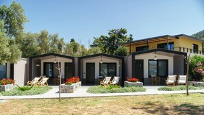 A modern three-unit residential building with a flat roof, large windows, and a covered outdoor seating area with lounge chairs and red flowers. - Via Lassi bungalow 6 (Ioanian Islands)