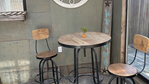 A rustic outdoor seating area with a wooden table and two stools, a large wall clock, and a 'WELCOME' sign.