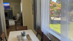 A dining area with a table set for four, a TV in the background, and a view of a garden through a window.