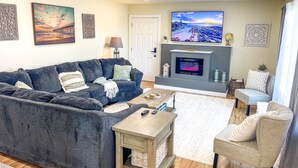 A living room with a dark grey sofa, a small table, a grey armchair, a fireplace with a TV, and decorative wall art.
