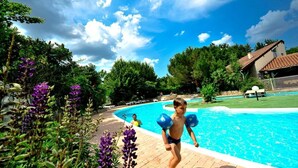 Une piscine sous un ciel dégagé, entourée de verdure et bordée d’un bâtiment dont le toit est recouvert de tuiles.