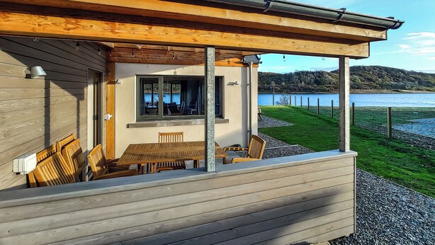 Een houten terras met een tafel en stoelen, met uitzicht op een meer en heuvels.