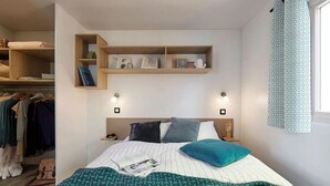 A bedroom with a bed, a wardrobe, a shelf with books and decorative items, and a window with a curtain.
