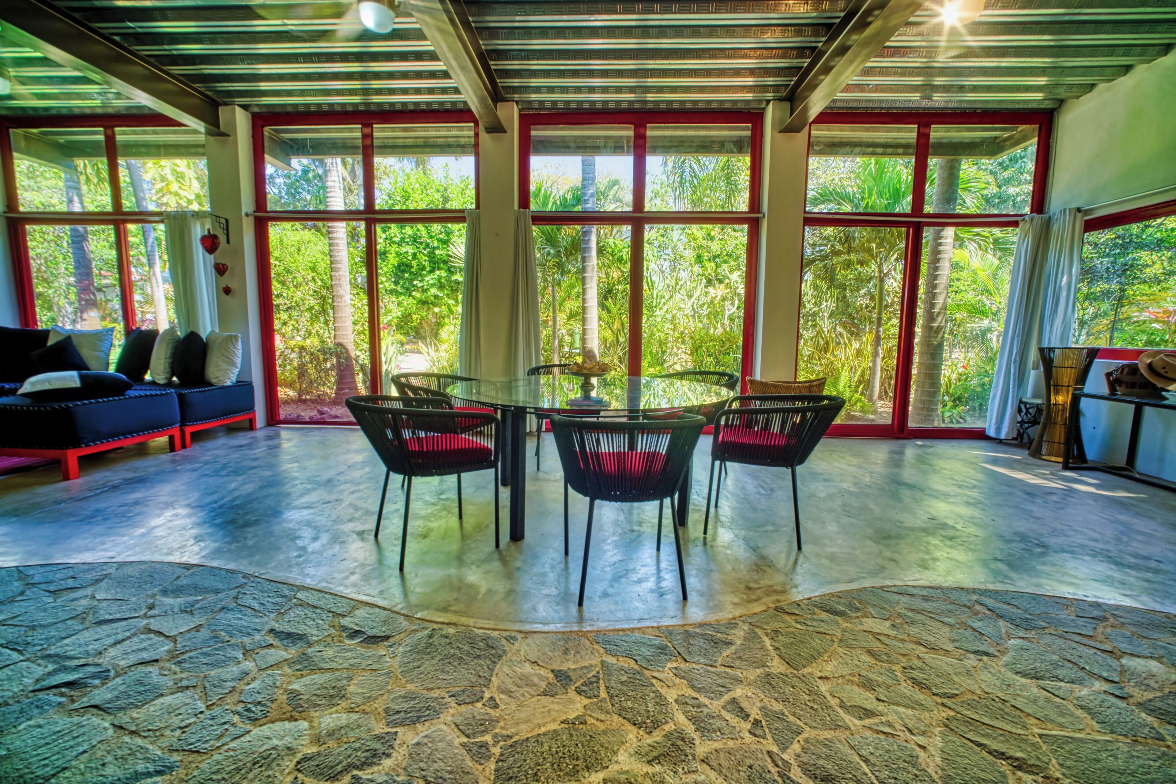 A spacious room with a stone floor, a glass table surrounded by chairs, and large windows offering a view of lush greenery.