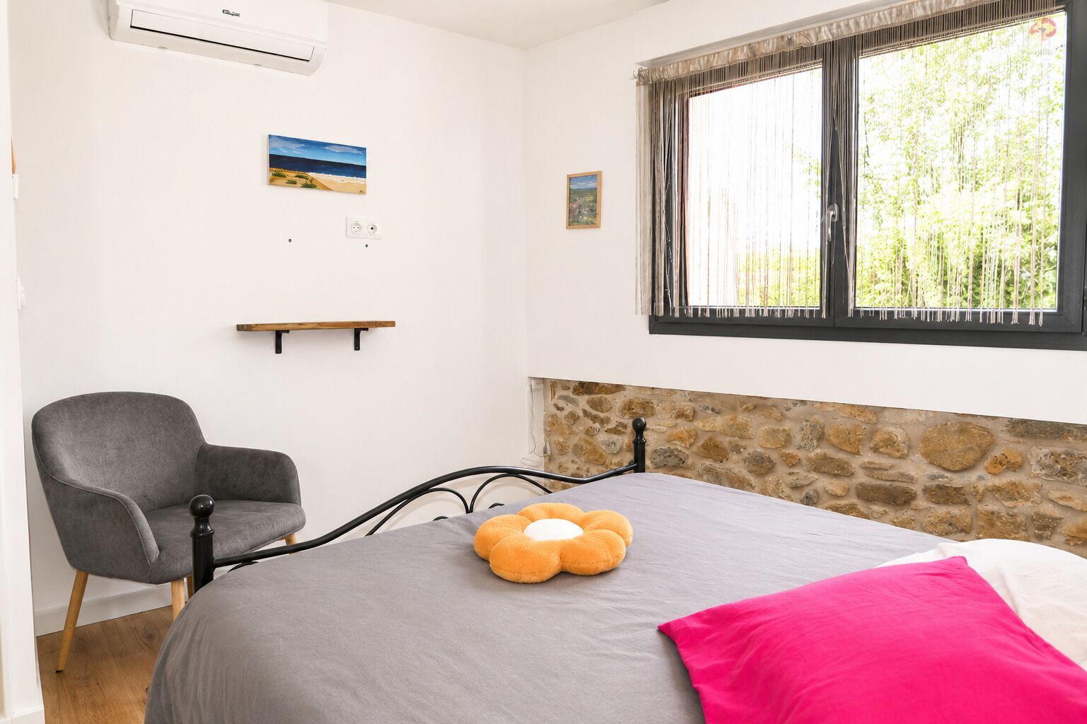 Ein Schlafzimmer mit einem Bett, einem Sessel, einem Fenster mit Vorhängen, einer Steinwand und einem kleinen Regal.