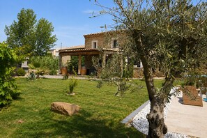 A stone villa with a tiled roof, a covered patio, and a well-maintained garden with a large rock, olive tree, and various plants.