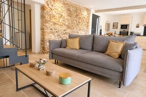 A living room with a stone wall, a grey sofa, a wooden coffee table, and a kitchen in the background.