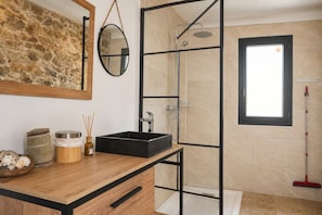 Un baño con tocador de madera, lavamanos negro, cabina de ducha con mampara de cristal y ventana con vistas.