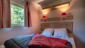 A bedroom with a bed, two bedside lamps, a window with curtains, and a wooden headboard. - ZEcamping 3 étoiles piscine chauffée locatif TV clim 21m2 4 pers (Soreze)