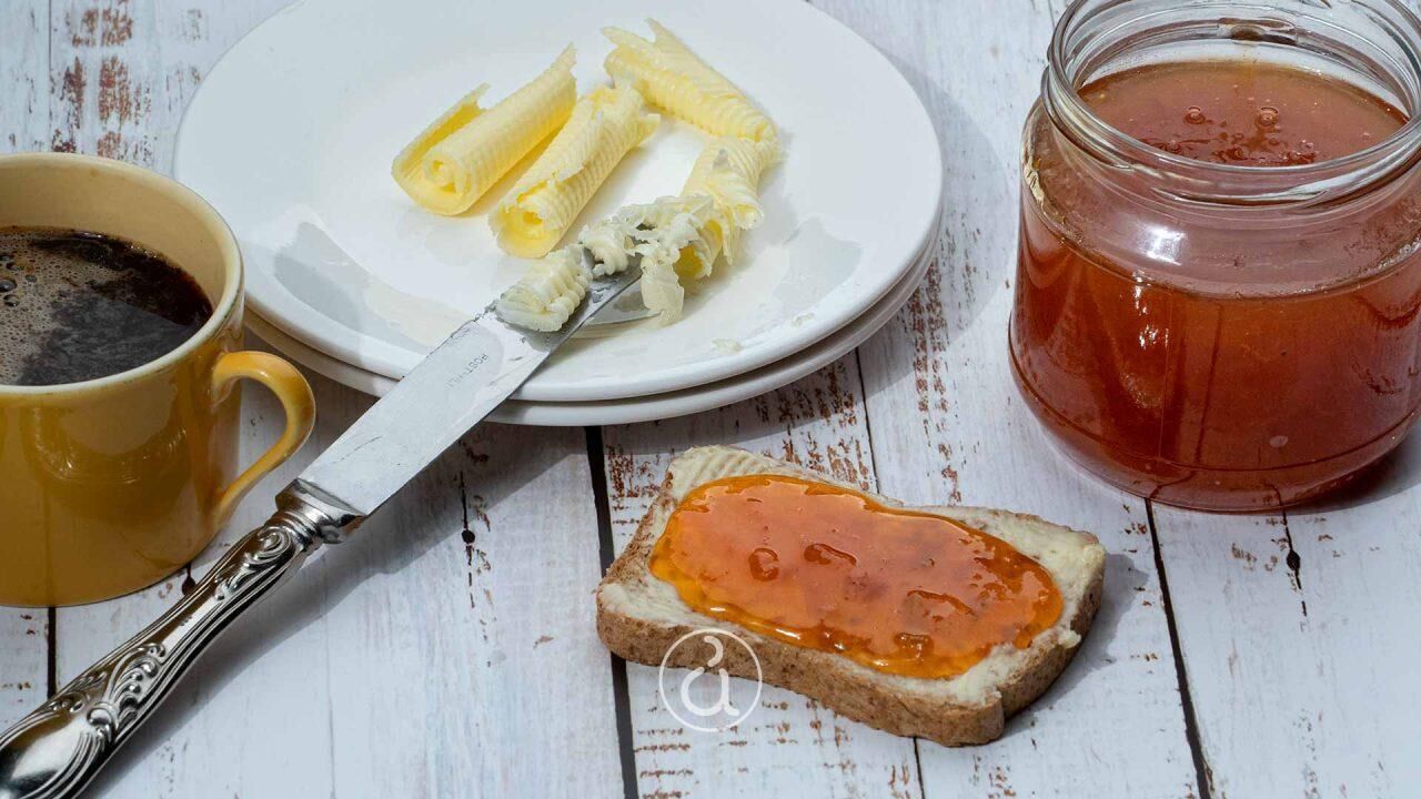 A plate with banana slices, a jar of jam, a slice of bread with jam, a cup of coffee, and a knife.