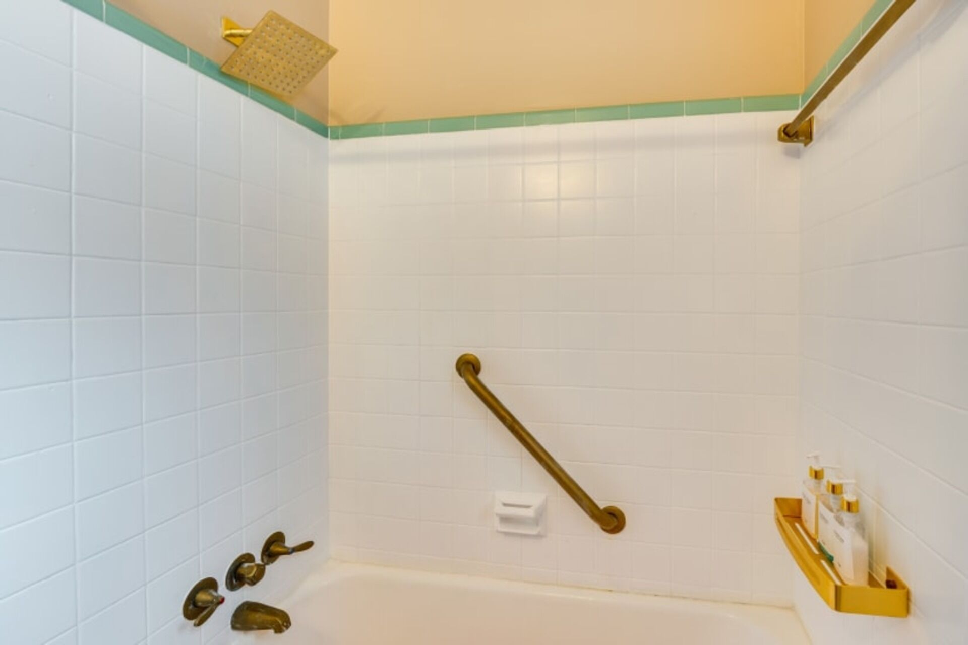 A bathroom with white tiled walls, a bathtub, a shower with a handheld showerhead, and a gold-colored grab bar.