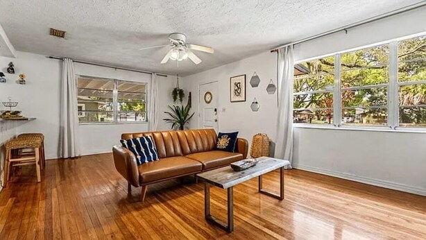 Un salon spacieux doté d’un plancher en bois, d’un canapé en cuir marron, d’un ventilateur de plafond et de grandes fenêtres.