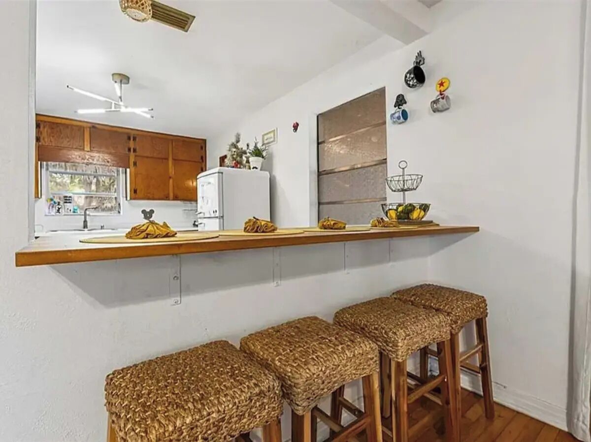 Cucina con piano di lavoro in legno, frigorifero e lavello. Quattro sgabelli di vimini e una ventola a soffitto.