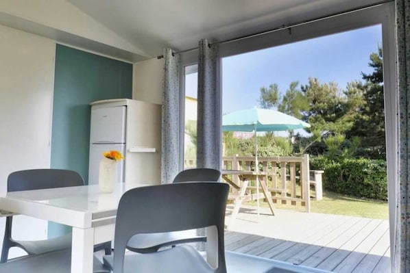 A balcony with a table and chairs, a refrigerator, and a view of a garden with a wooden deck and a green umbrella.
