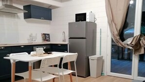 Une cuisine moderne avec des meubles blancs, un réfrigérateur en acier inoxydable, une table à manger avec des chaises blanches et une porte-fenêtre coulissante avec des rideaux.