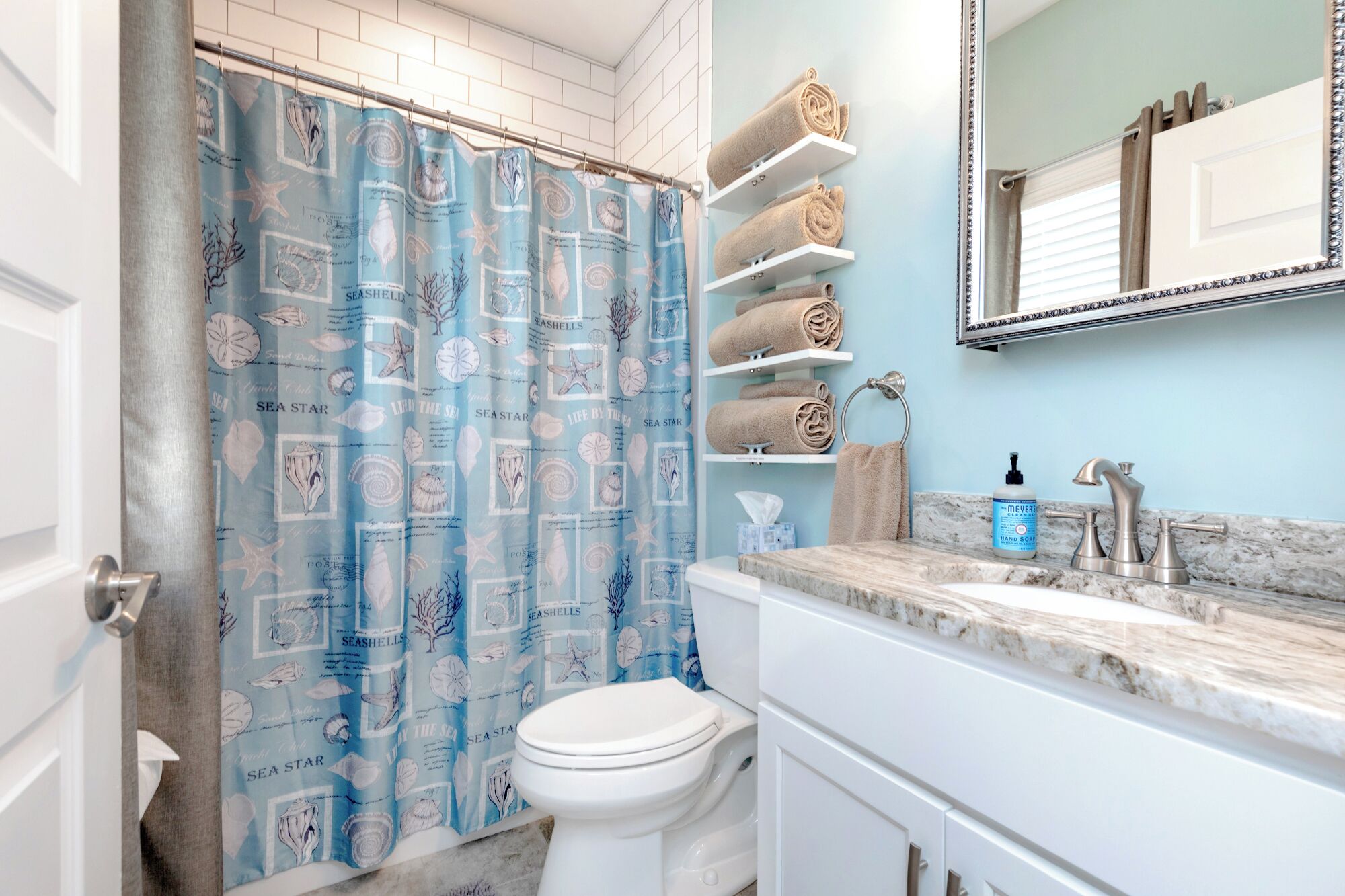 A bathroom with a blue seashore-themed shower curtain, white toilet, marble sink, and shelves with rolled towels.