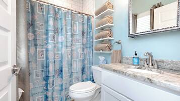 A bathroom with a blue seashore-themed shower curtain, white toilet, marble sink, and shelves with rolled towels.