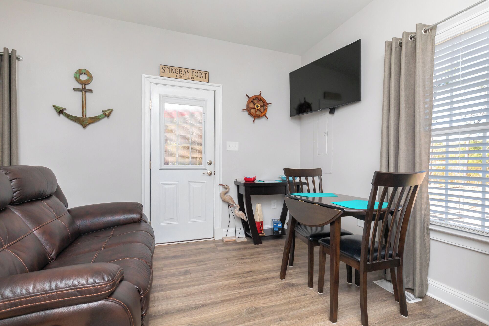 A living room with a leather couch, a dining table with chairs, a TV mounted on the wall, and a sign above the door.