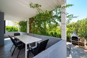 An outdoor patio with a white table and black chairs, a stone barbecue grill, and lush greenery in the background. - Bbq Holly Garden Novalja D (Novalja)