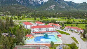 A hotel with multiple red-roofed buildings, a swimming pool, tennis courts, and a mountainous backdrop.