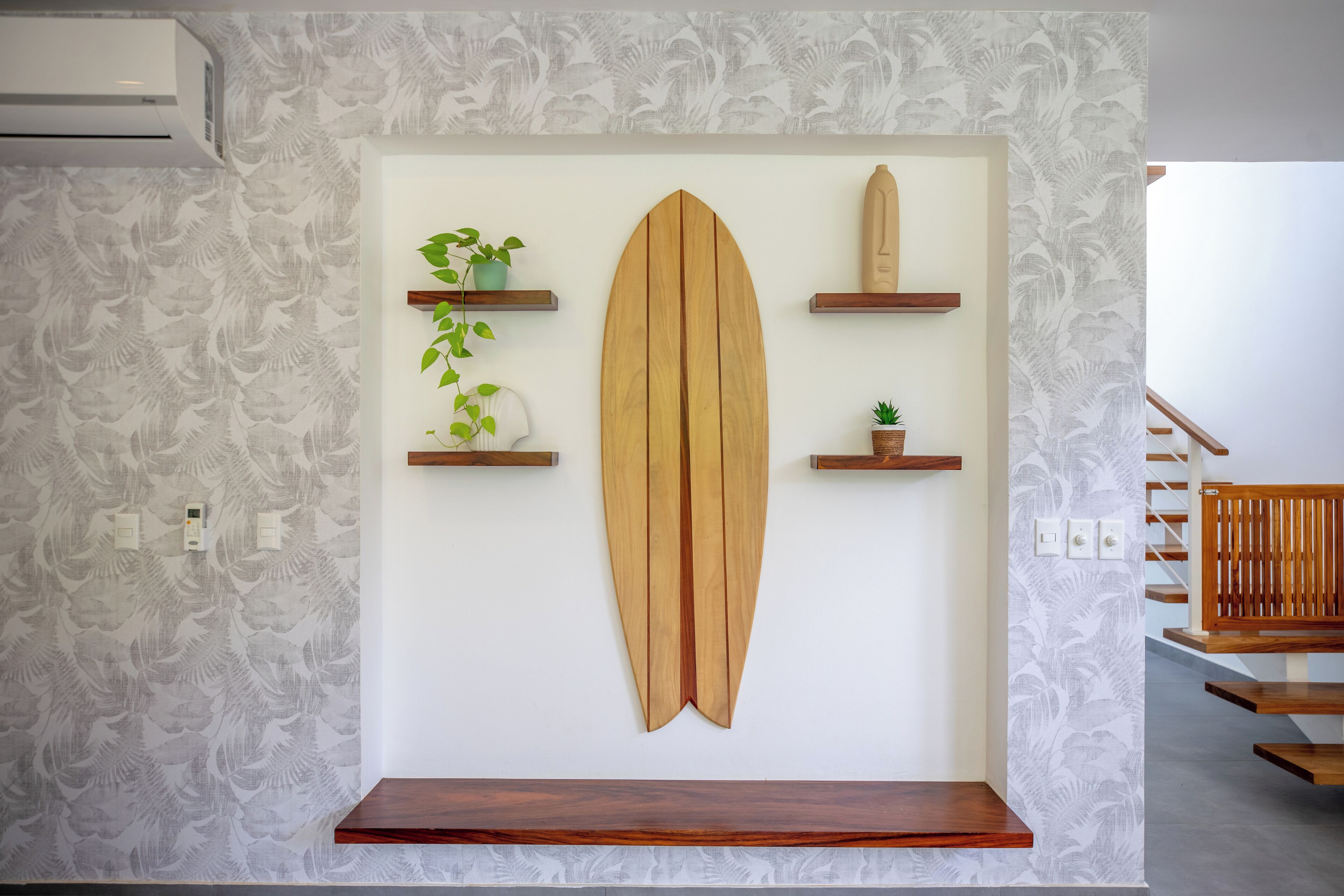 Una pared con una tabla de surf, tres estantes de madera con plantas y un banco de madera.