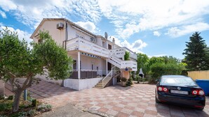 A two-story building with a checkered driveway, a car parked in front, and a tree in the foreground. - Apartments Valy 6 Center Novalja (Novalja)