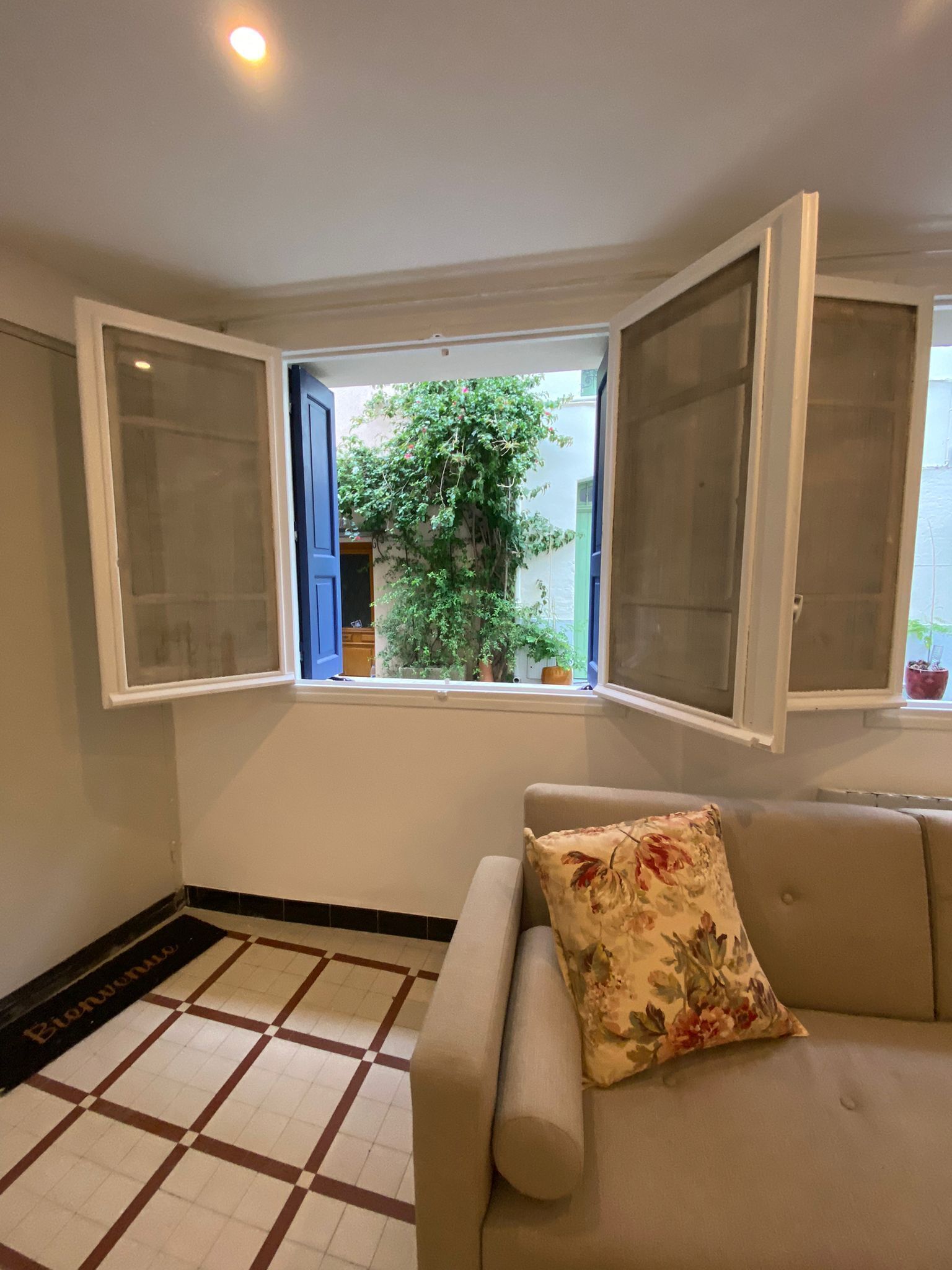 A room with a beige sofa, a floral pillow, a window with open shutters, and a tiled floor.
