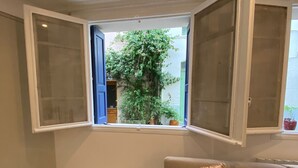 A room with a beige sofa, a floral pillow, a window with open shutters, and a tiled floor.