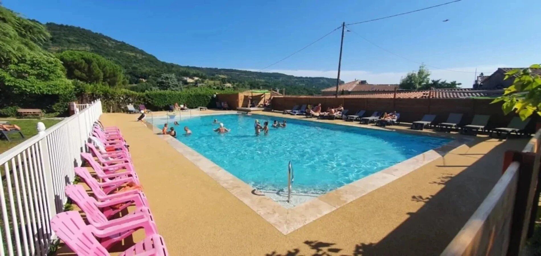 A swimming pool with people in the water and on the pool deck, surrounded by lounge chairs and a white fence.
