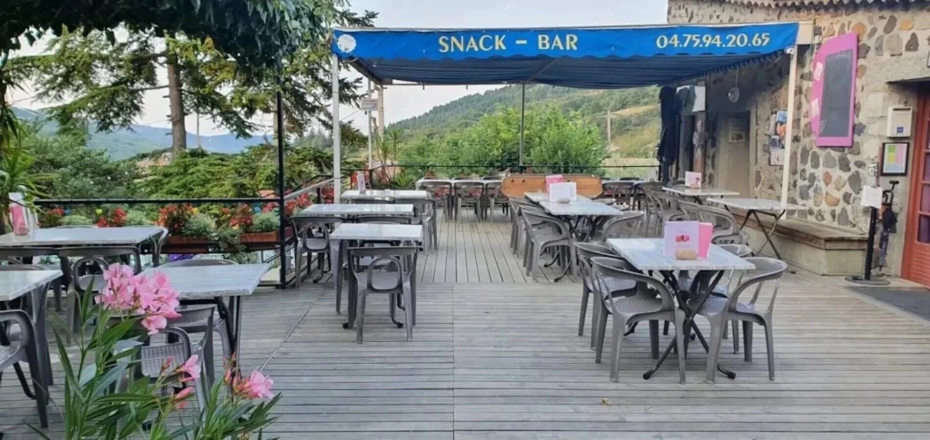 Un área de comedor al aire libre con mesas y sillas, un toldo azul con la leyenda 'SNACK - BAR' y un número de teléfono, y un edificio de piedra al fondo.