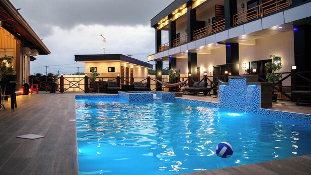 Una piscina con una hamaca y sillas de descanso, rodeada por un edificio con varios balcones e iluminación exterior.