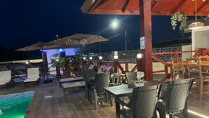 Un espacio de comedor al aire libre con mesas y sillas, piscina y luces colgantes.