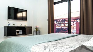 A hotel room with a bed, a TV mounted on the wall, a small table with a vase of flowers, and a view of an outdoor area through a large window. - Yasmin Luxury Resort (Paramaribo)